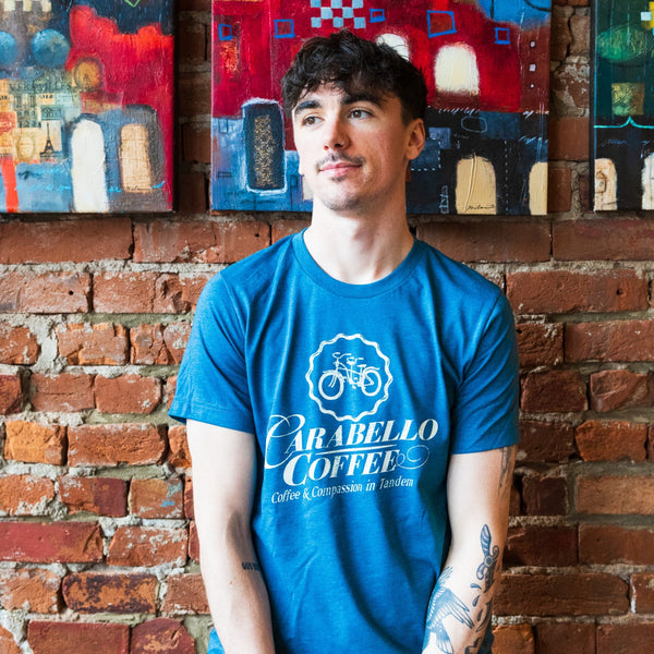Man wearing teal Carabello Coffee logo tee shirt in front of a brick wall 