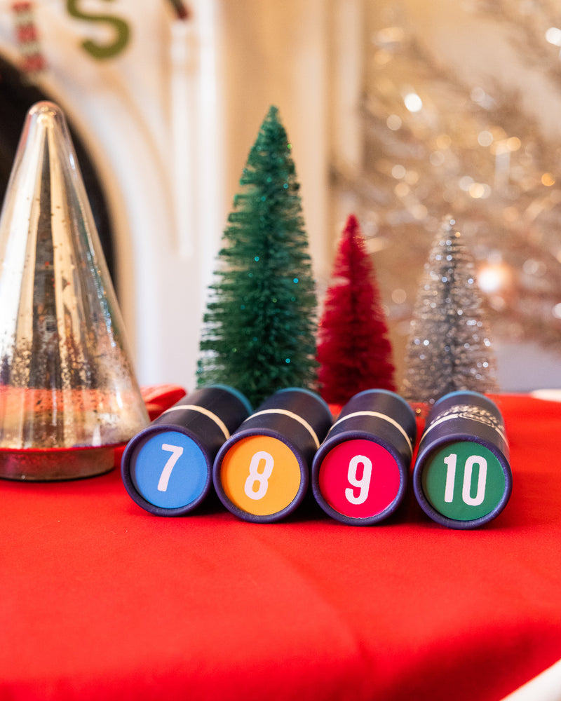 12 Days of Christmas coffee with numbered tubes on a red surface, Christmas trees in the background