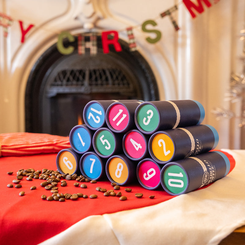Stack of colorful numbered canisters on a table with coffee beans, Christmas decorations in the background.