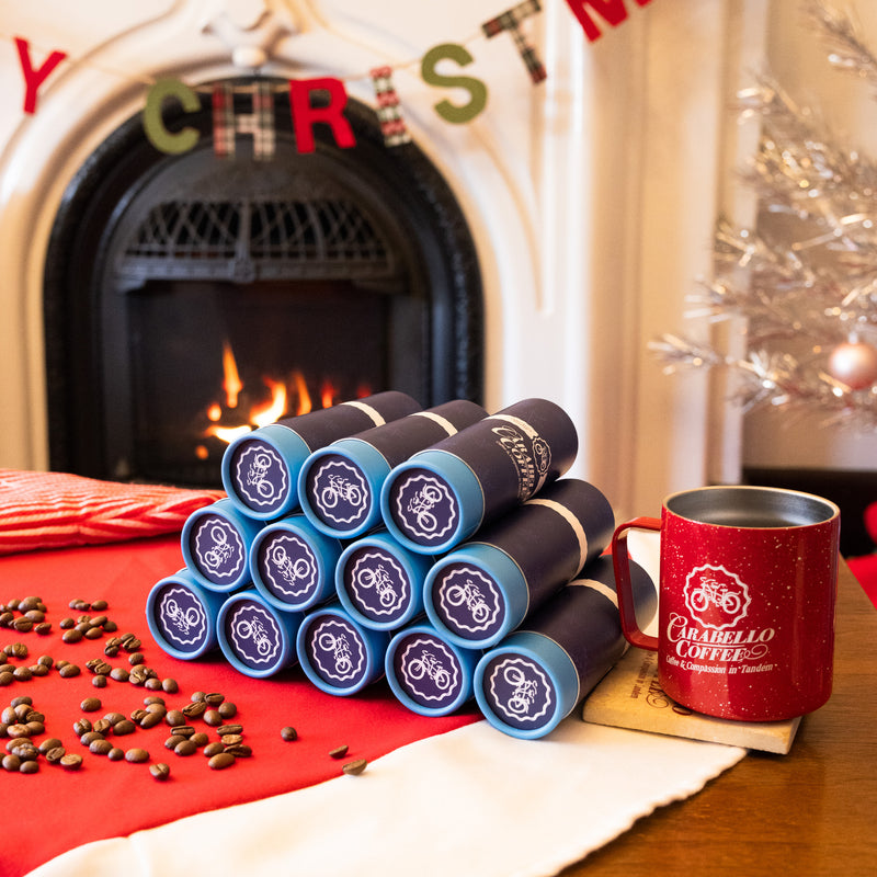 Stack of Caribello Coffee packages with a red mug in front of a fireplace with Christmas decorations.