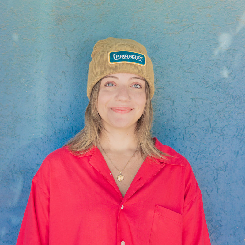 Person wearing a beige beanie with a logo and a red shirt against a blue wall