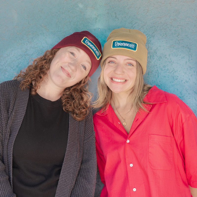 Two people wearing beanies with visible branding against a blue textured wall.