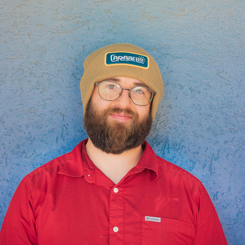 Man wearing a beige beanie with a logo and a red shirt against a blue textured wall