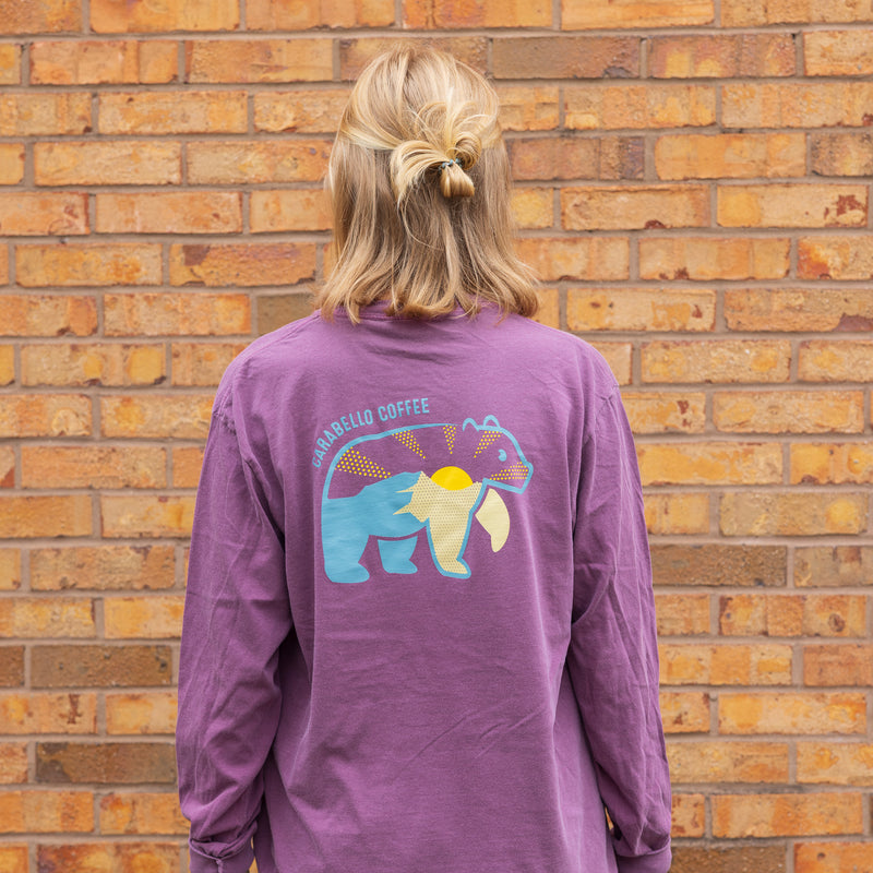 Person wearing a purple long-sleeve shirt with a bear graphic against a brick wall.