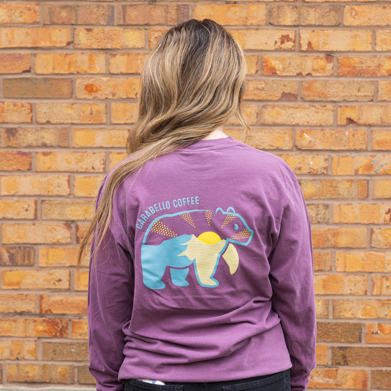 Person wearing a purple long-sleeve shirt with a bear graphic and text, standing against a brick wall.