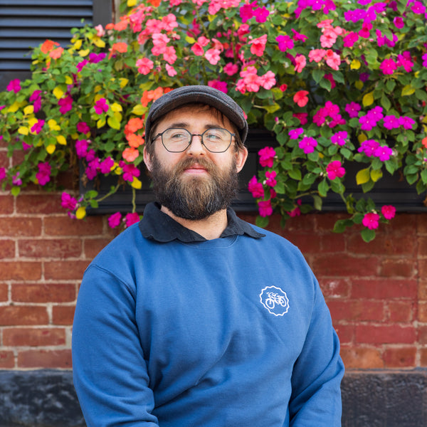 Man with a beard wearing a blue sweater with a logo in front of colorful flowers and a brick wall.