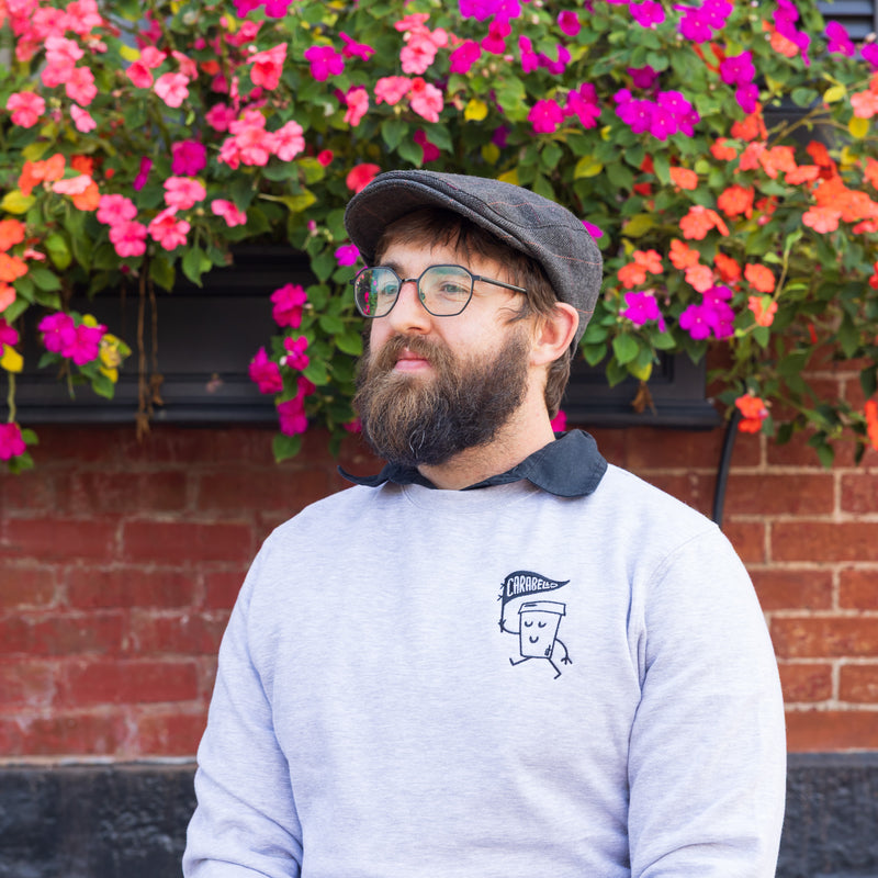 Man with a beard wearing a flat cap and glasses, standing in front of colorful flowers.
