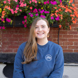Woman wearing a blue sweatshirt with a logo in front of colorful flowers and a brick wall.