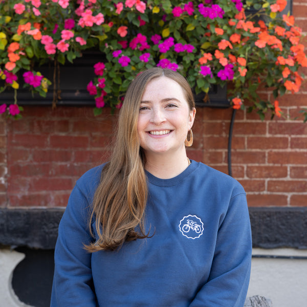 Woman wearing a blue sweatshirt with a logo in front of colorful flowers and a brick wall.