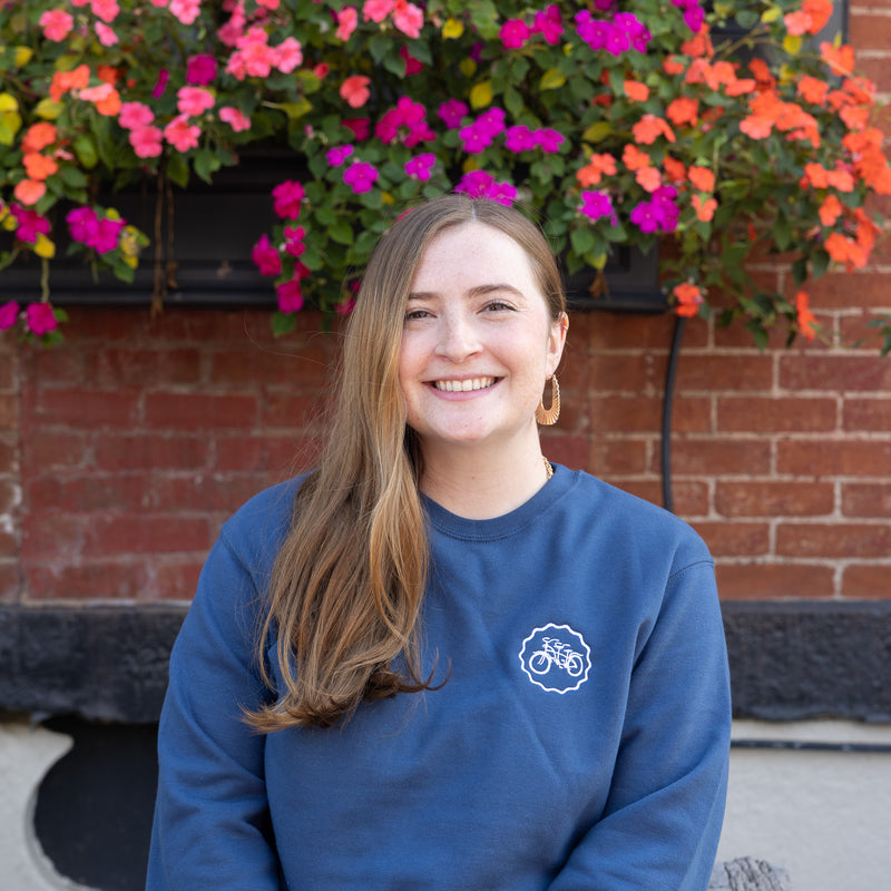 Woman wearing a blue sweatshirt with a logo in front of colorful flowers and a brick wall.