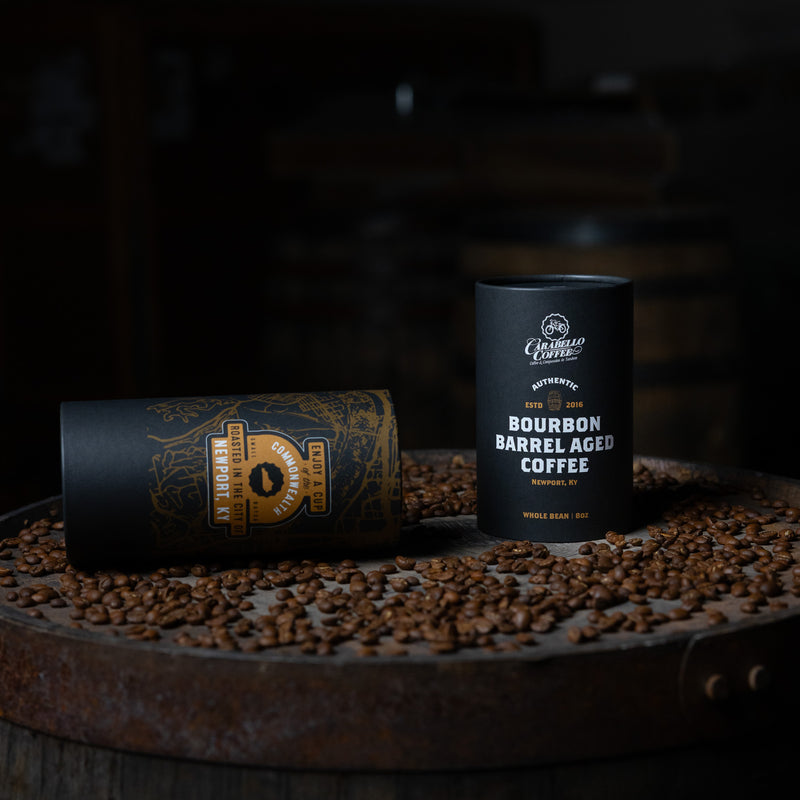 Two coffee containers labeled 'Bourbon Barrel Aged Coffee' on a bed of coffee beans with a dark background.