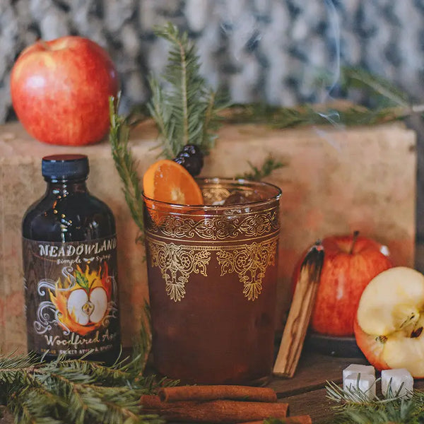 Meadowland syrup bottle with a glass of dark liquid, apples, and cinnamon sticks on a wooden surface.