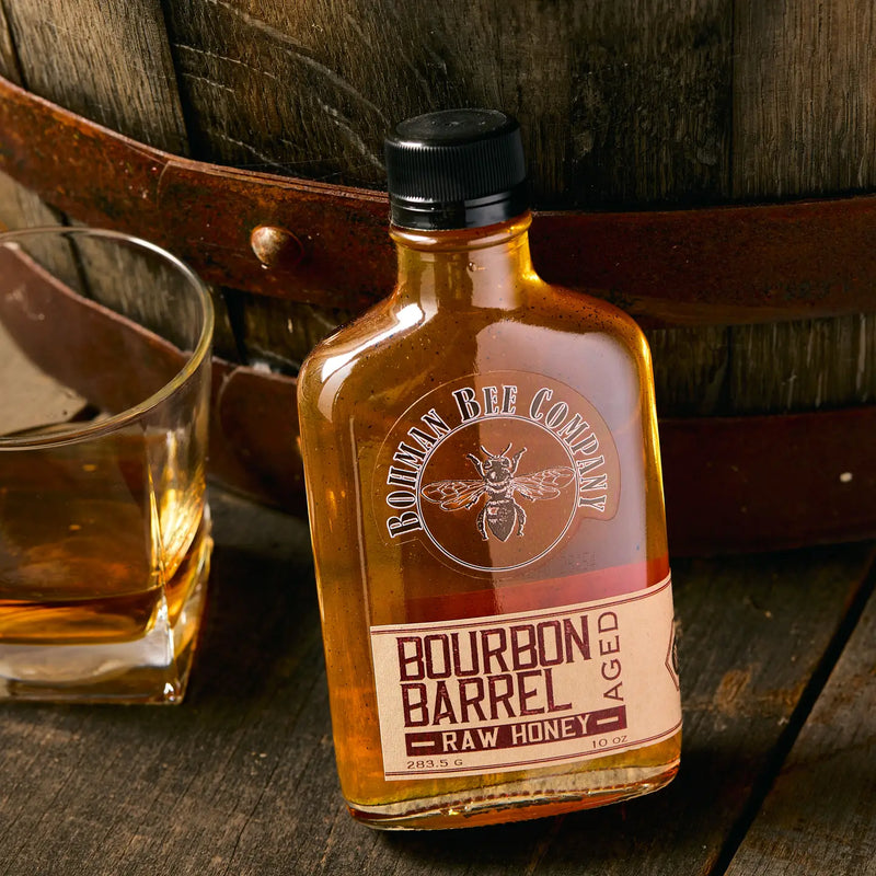 Bottle of Bourbon Barrel Raw Honey with a glass and barrel in the background