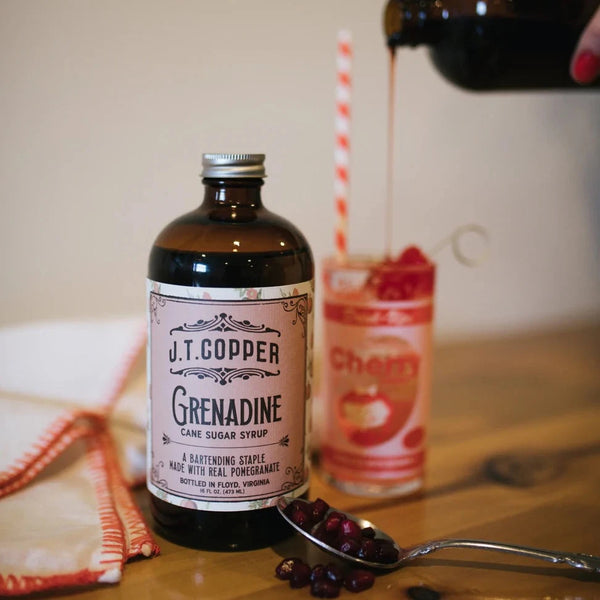 8 oz bottle of grenadine syrup with a cocktail being mixed in the background.  A spoon filled with pomegranate seeds in the front.  