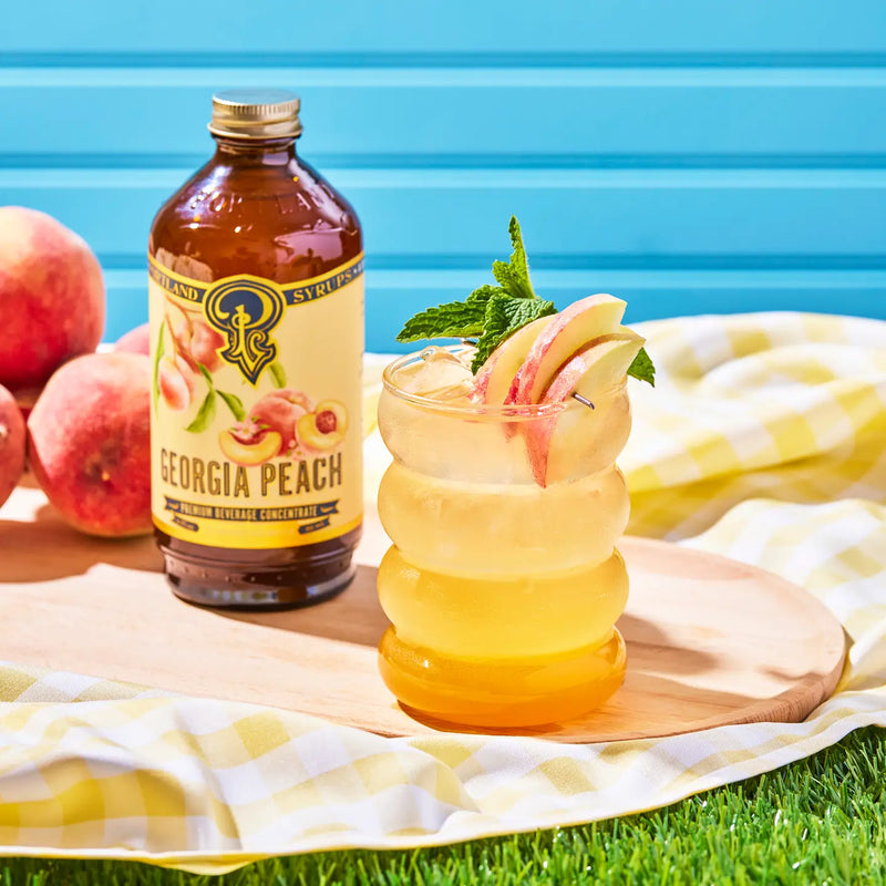 Glass bottle of peach syrup next to iced tea topped with peaches and mint leaves
