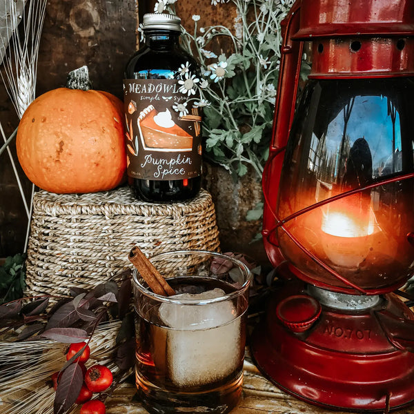 Meadowland Pumpkin Spice simple syrup bottle with a lit lantern, glass of liquid, and pumpkin on a rustic background.